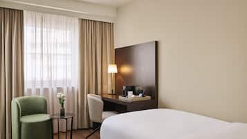 Habitación de hotel con cama, escritorio con lámpara, silla y una mesita con un jarrón de flores.