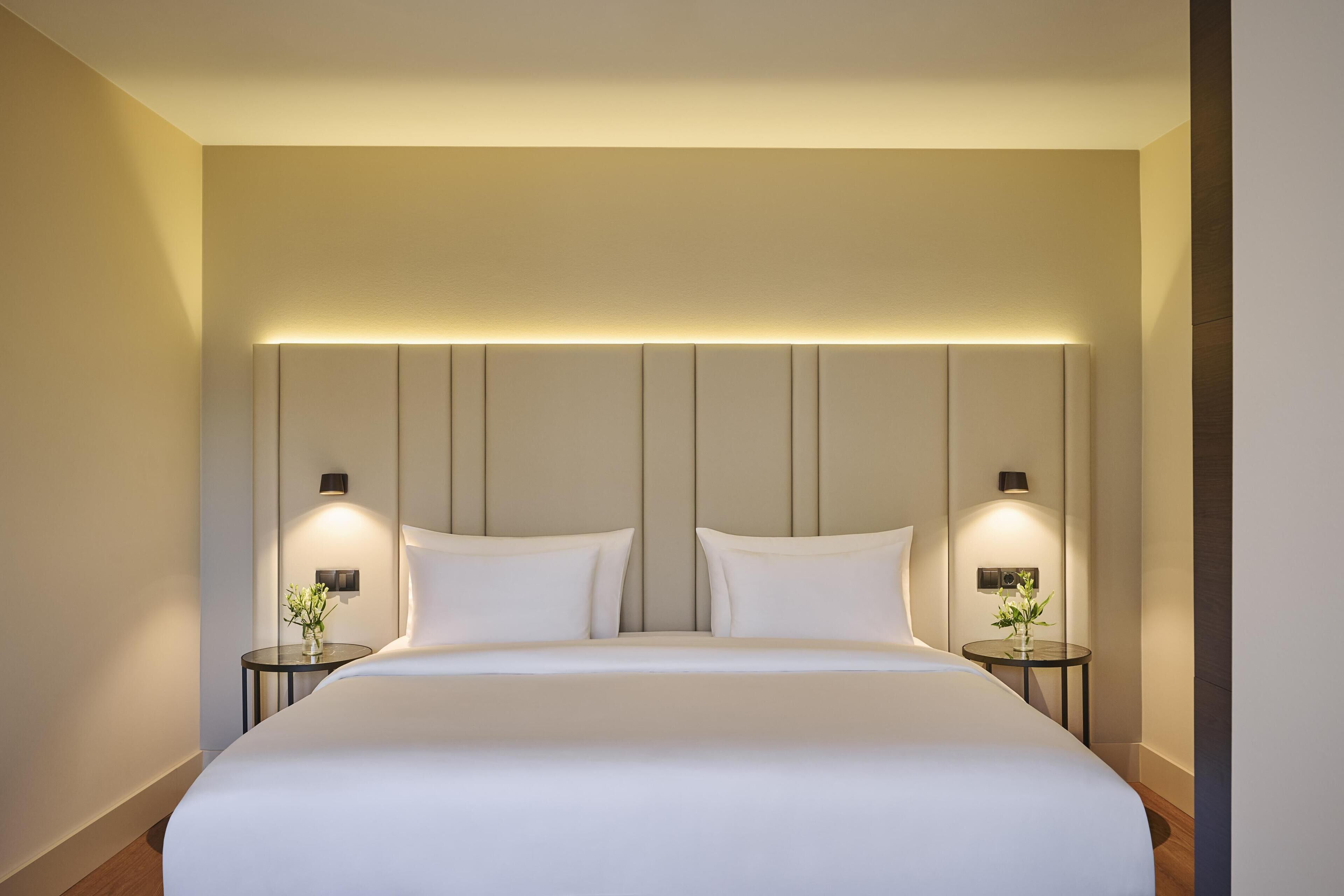 A modern hotel room with a large bed, two bedside tables with lamps, and a wall-mounted light fixture above the bed.
