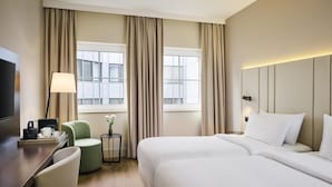 Camera d'albergo con due letti, una scrivania, una sedia, una lampada e vista sugli edifici attraverso ampie finestre.