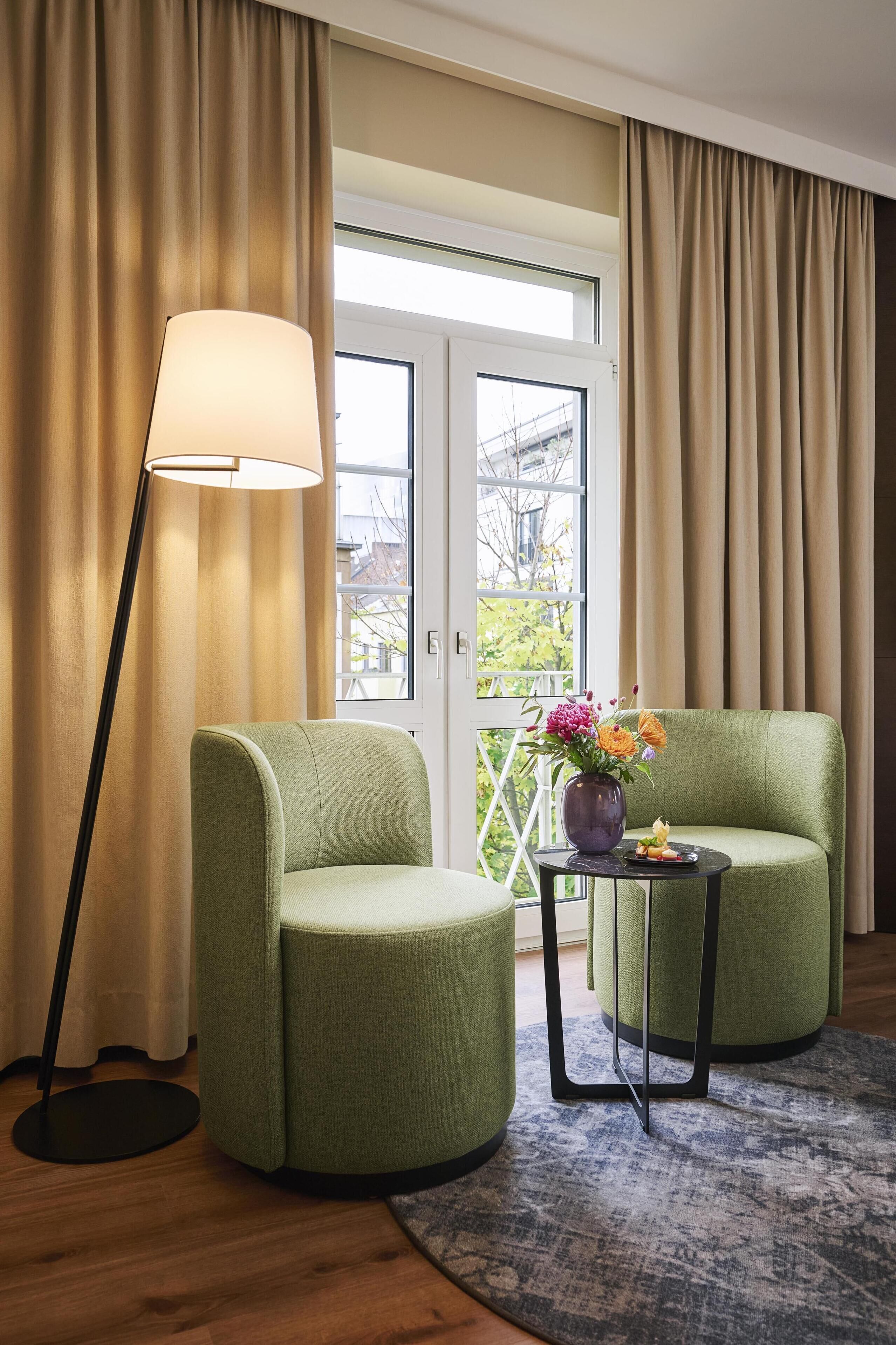 A room with two green armchairs, a floor lamp, a small table with a vase of flowers, and a view through a glass door.
