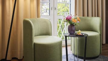 A room with two green armchairs, a floor lamp, a small table with a vase of flowers, and a view through a glass door.