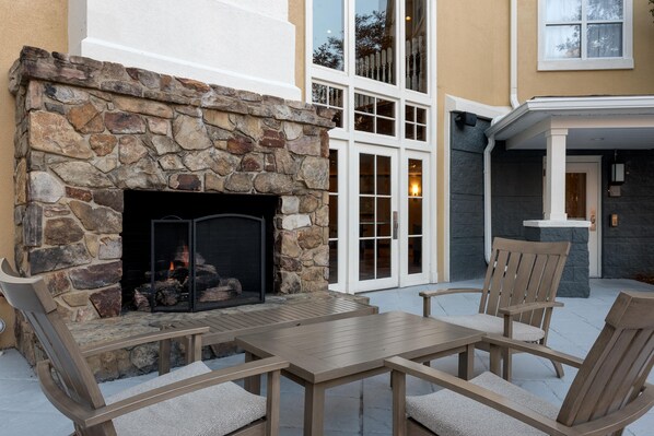 A stone fireplace with a fire, outdoor seating, and a building with large windows and a balcony.