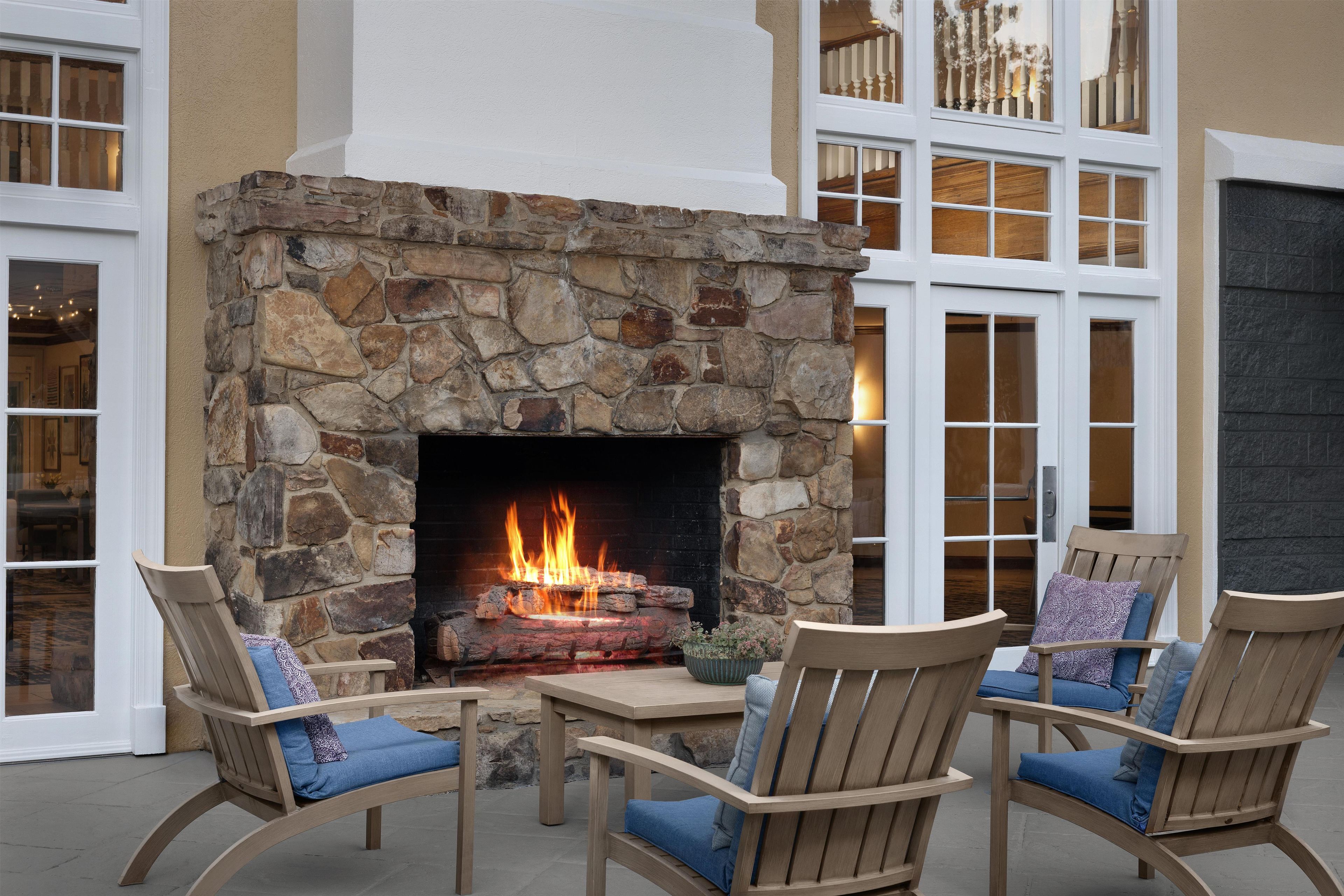 A stone fireplace with a fire, surrounded by wooden chairs with blue cushions.