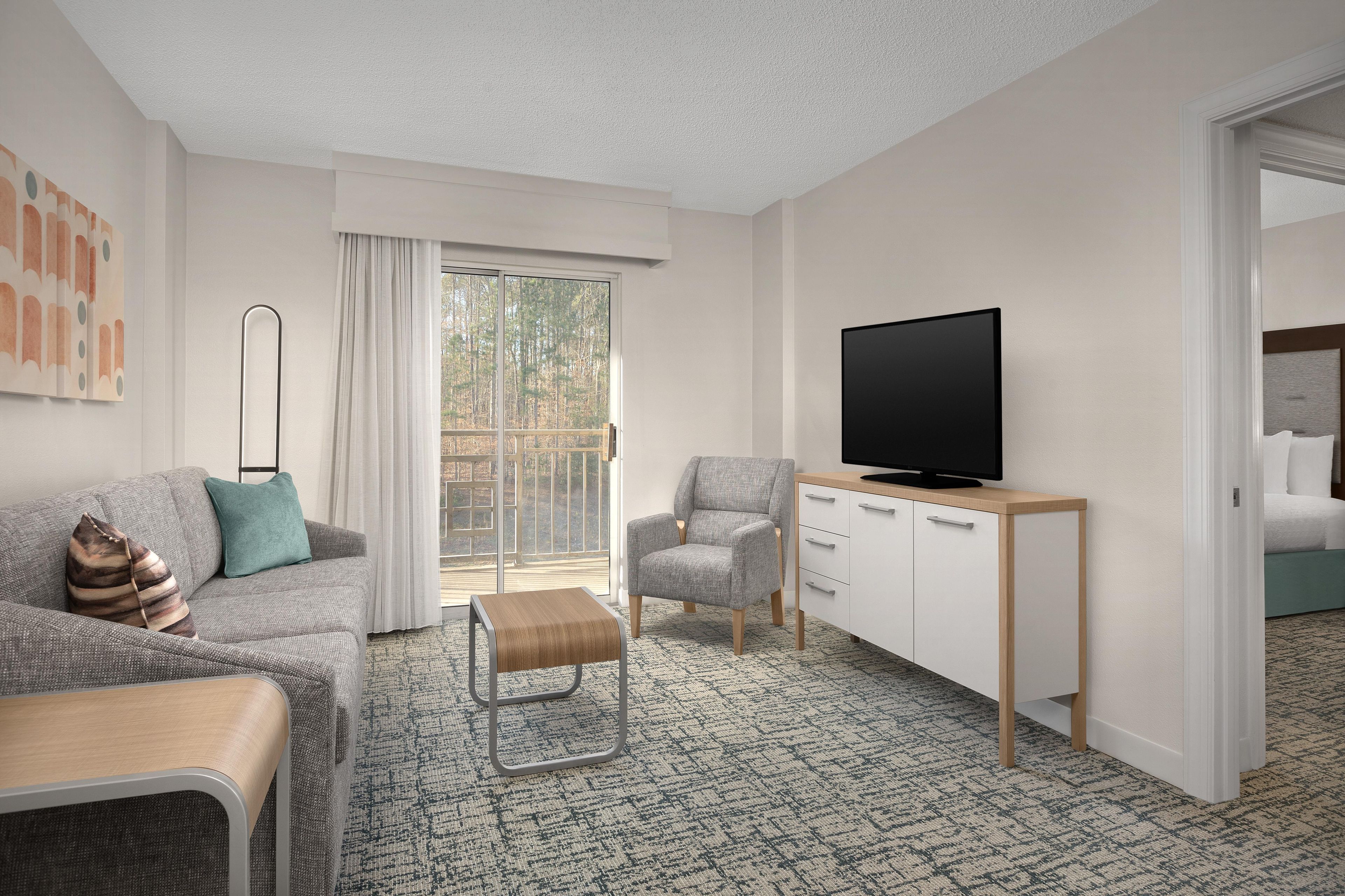 A modern hotel room with a grey sofa, a small wooden table, a grey armchair, a TV on a wooden stand, and a large window with a view of trees.