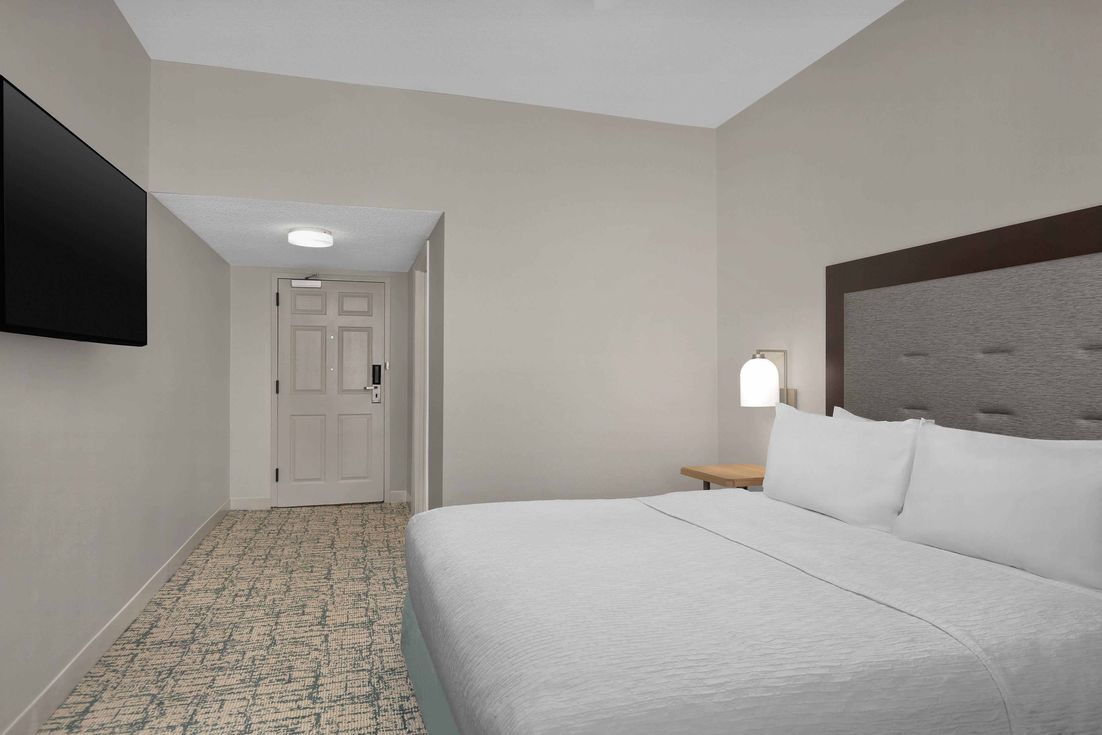 A hotel room with a bed, a flat-screen TV mounted on the wall, a bedside table with a lamp, and a door leading to another room.