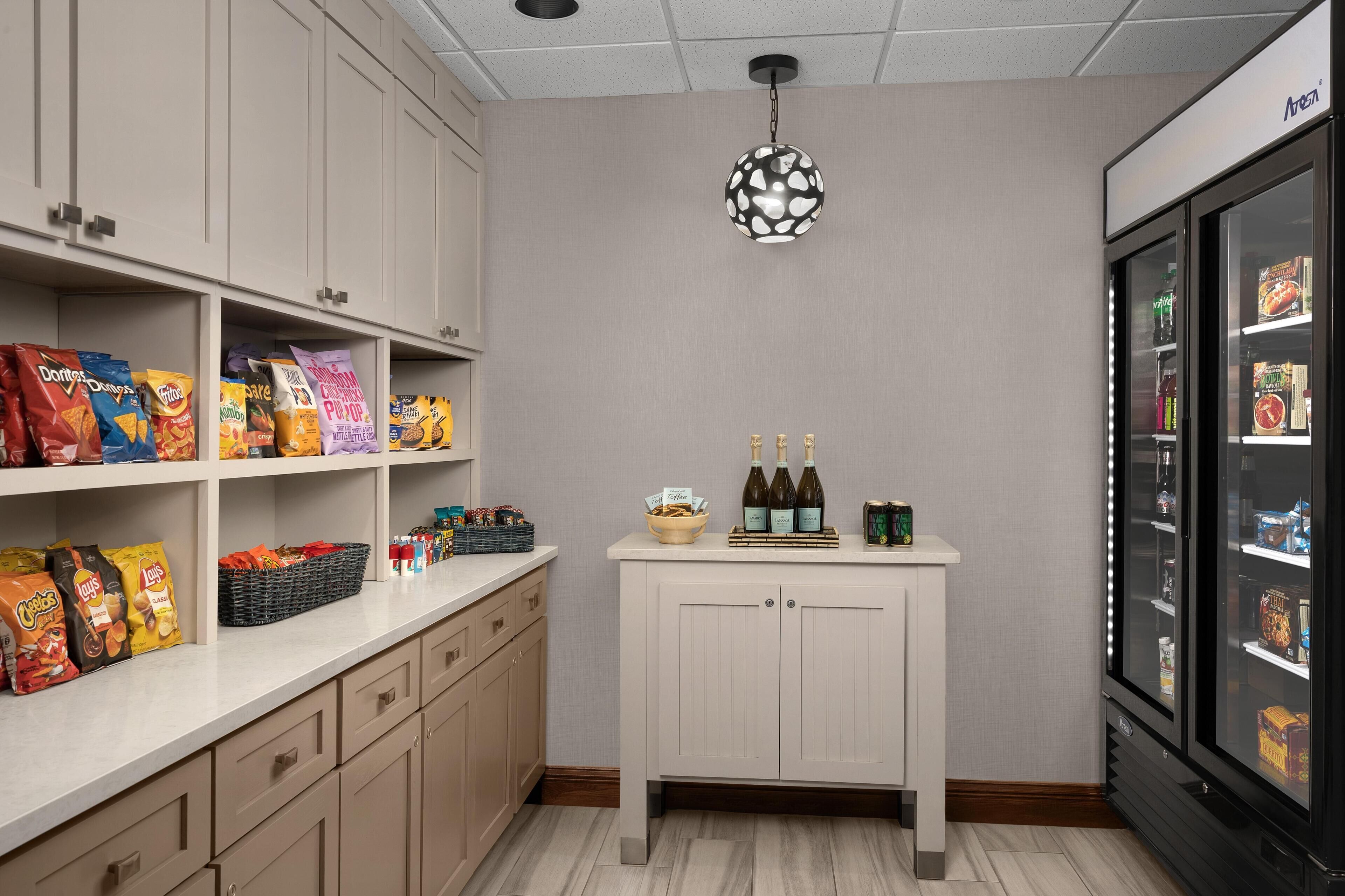 A kitchenette with shelves stocked with snacks, a small table with bottles, and a refrigerator.