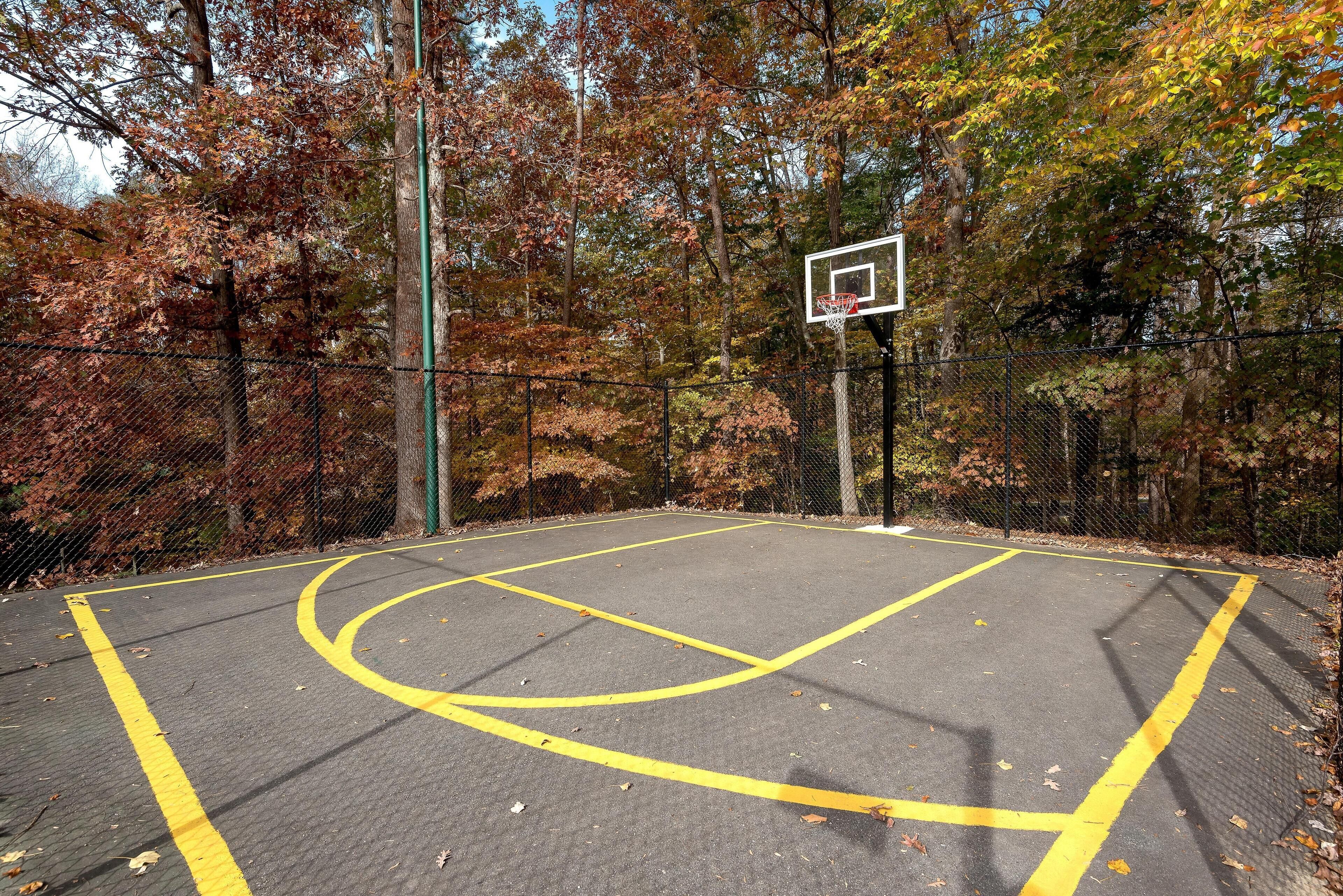 An outdoor basketball court with a chain-link fence, surrounded by trees with autumn foliage.