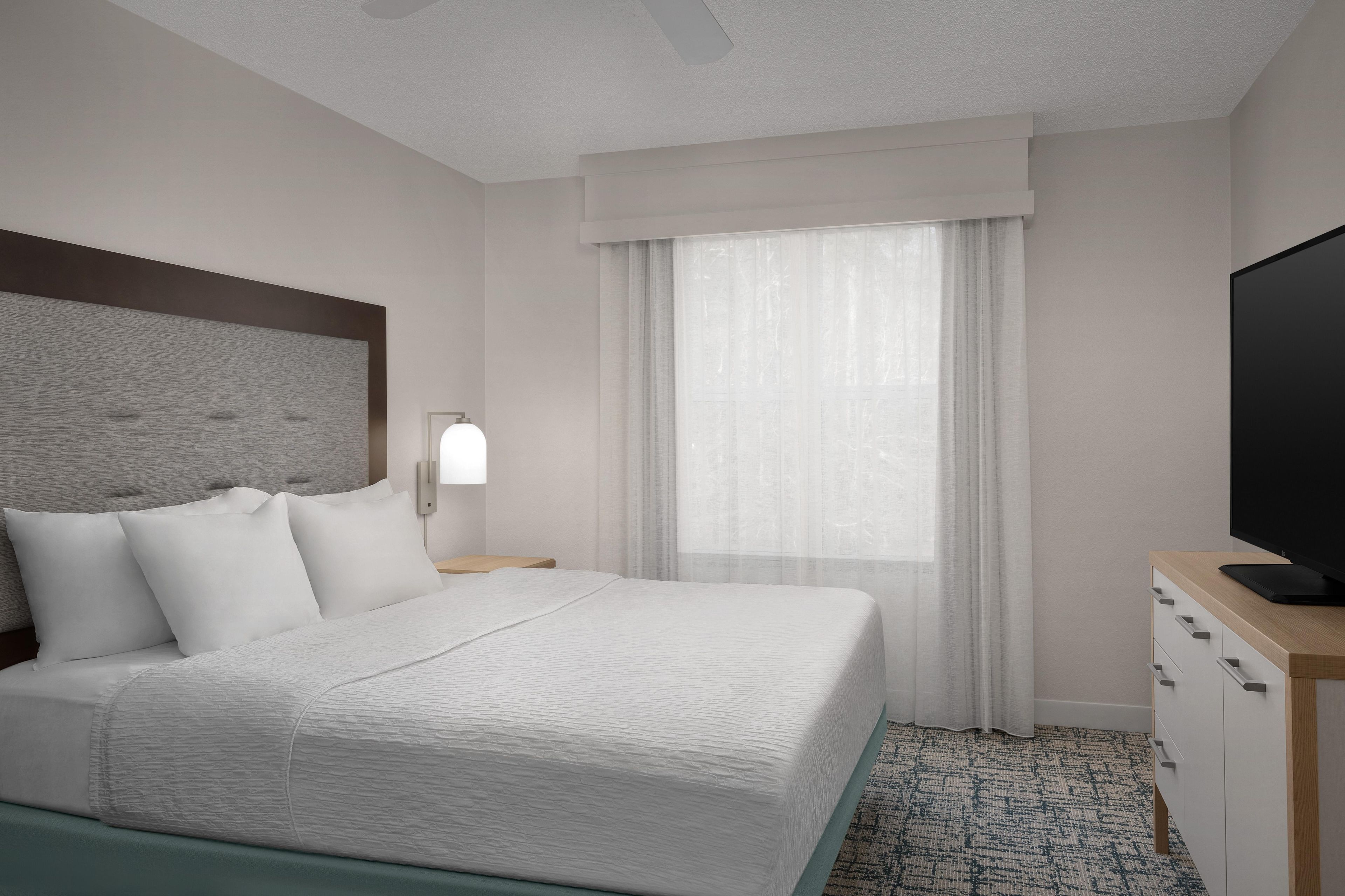 Una habitación de hotel con una cama, televisión, mesita de noche y ventana con cortinas.