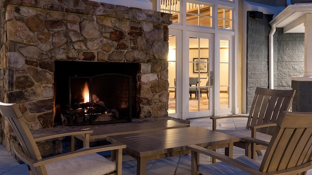 A stone fireplace with a fire, wooden furniture, and a view of a balcony with glass doors. - Homewood Suites by Hilton Raleigh/Cary (Cary)