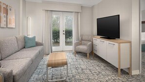 A modern hotel room with a grey sofa, a wooden coffee table, a grey armchair, a TV on a wooden stand, and a large window with curtains. - Homewood Suites by Hilton Raleigh/Cary (Cary)