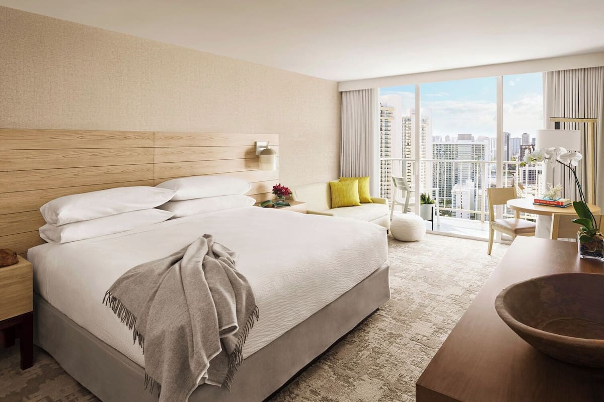 Habitación de hotel moderna con una cama grande, una mesita de noche con lámpara, un pequeño rincón de descanso con una silla, una mesa redonda con un cuenco, y vistas a la ciudad a través de amplios ventanales.