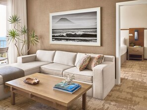 A living room with a beige sofa, a wooden coffee table, a framed picture of a mountain and ocean, and a view of the sea through a window.