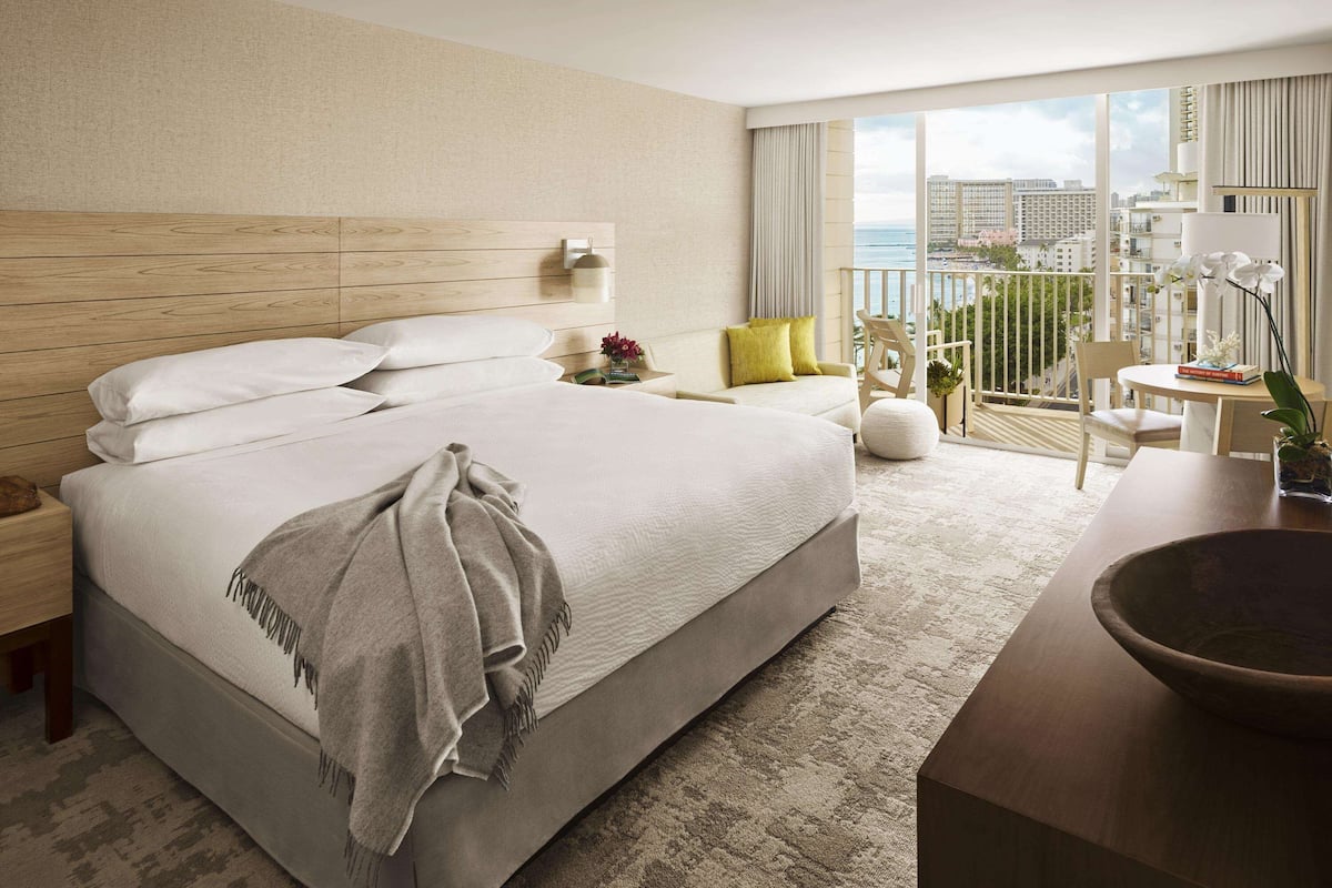 Habitación de hotel con una cama grande, un balcón con vistas, una mesita con un florero y una silla.