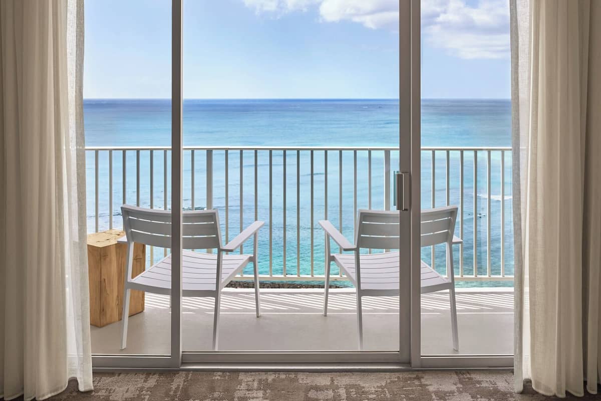 Un balcón con dos sillas blancas y una mesa de madera, con vista a un mar azul claro y una costa lejana.