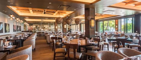 A spacious restaurant with high ceilings, wooden beams, and large windows. Tables are arranged in a casual layout, and there are hanging spherical light fixtures.
