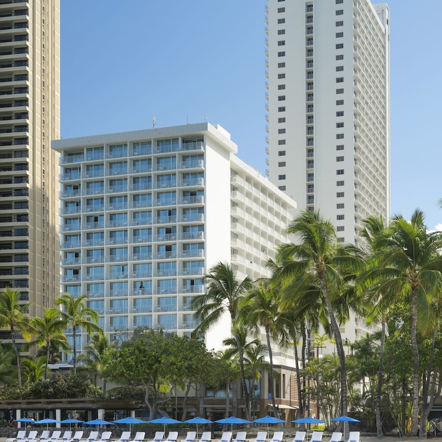 A beachfront hotel with multiple floors, a pool area with lounge chairs and umbrellas, and palm trees.