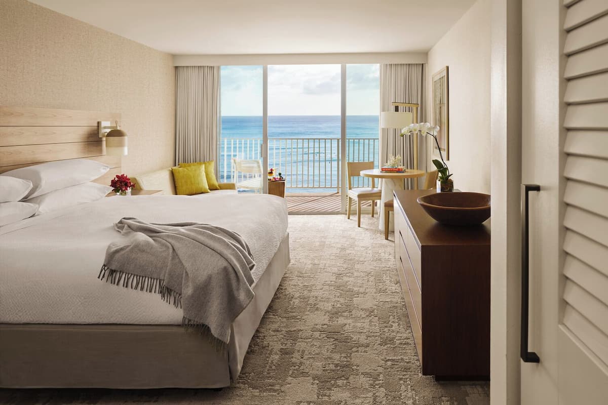 Habitación de hotel con una cama grande, un escritorio, una silla, una mesita y un frutero en el escritorio, con vista al océano a través de las puertas del balcón.