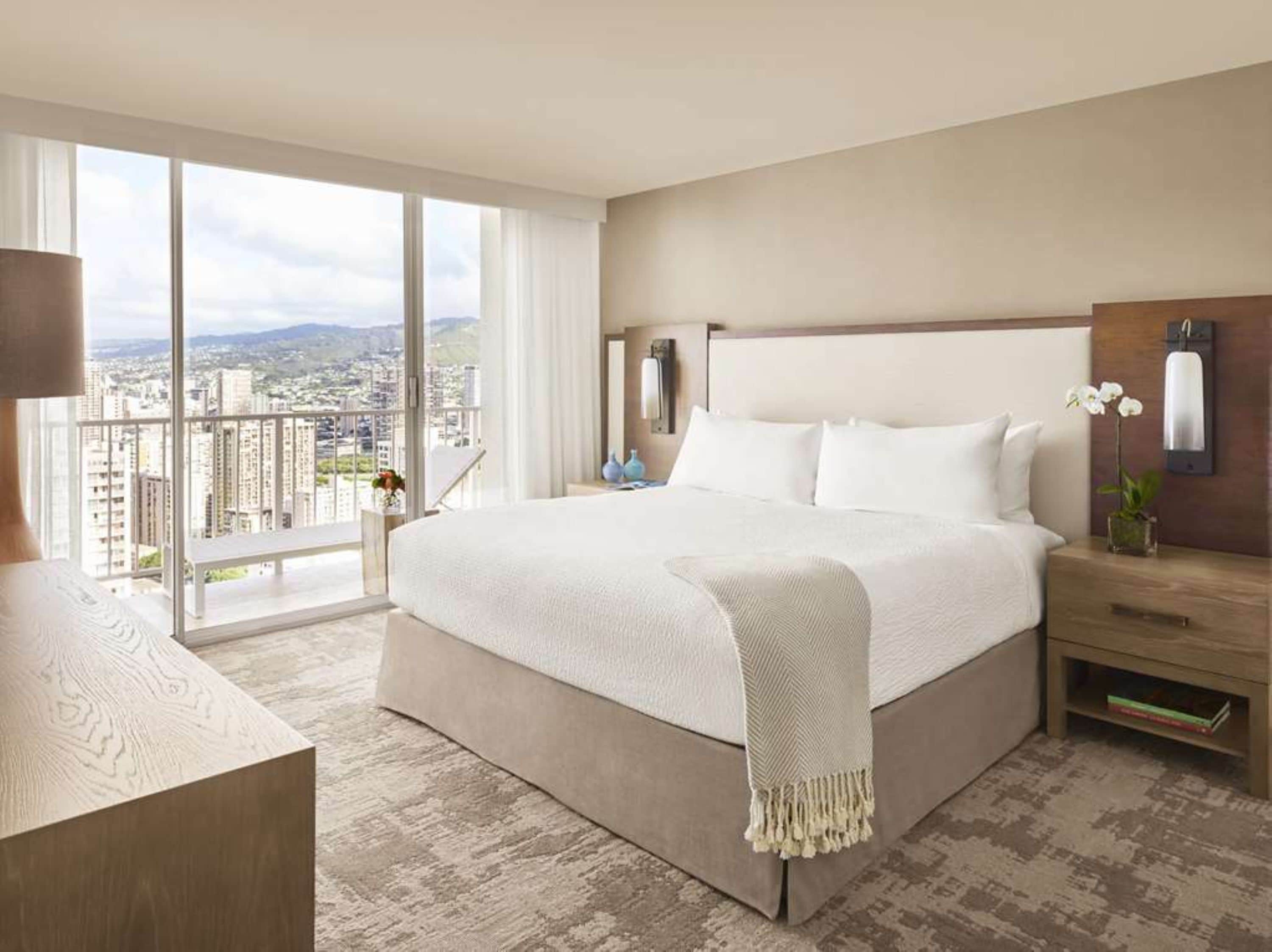 Une chambre d’hôtel avec un grand lit, une table de chevet, une lampe, un vase de fleurs et une vue sur la ville grâce à de grandes fenêtres.