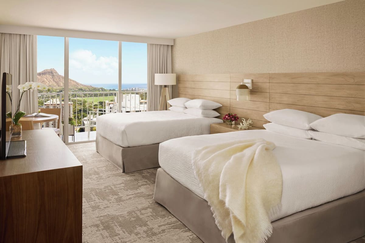 Habitación de hotel con dos camas, un escritorio y vistas a un paisaje montañoso.