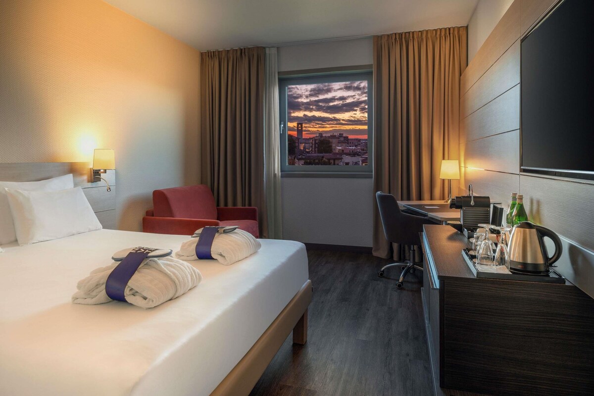 A hotel room with a bed, a desk, a chair, a window with a sunset view, and a TV on the wall.
