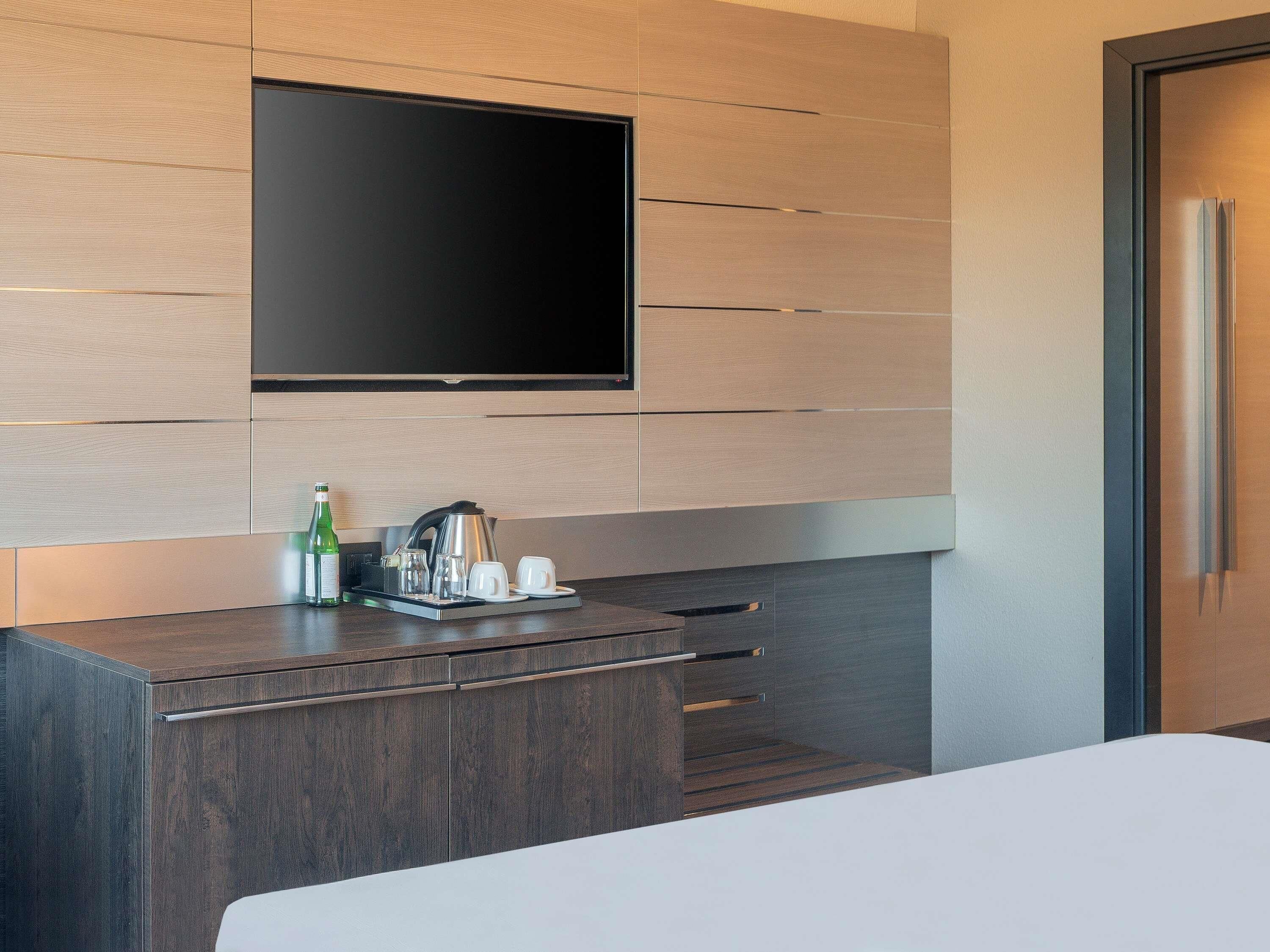 A modern hotel room with a flat-screen TV mounted on the wall, a minibar with a kettle, glasses, and a bottle, and a wooden cabinet with drawers.