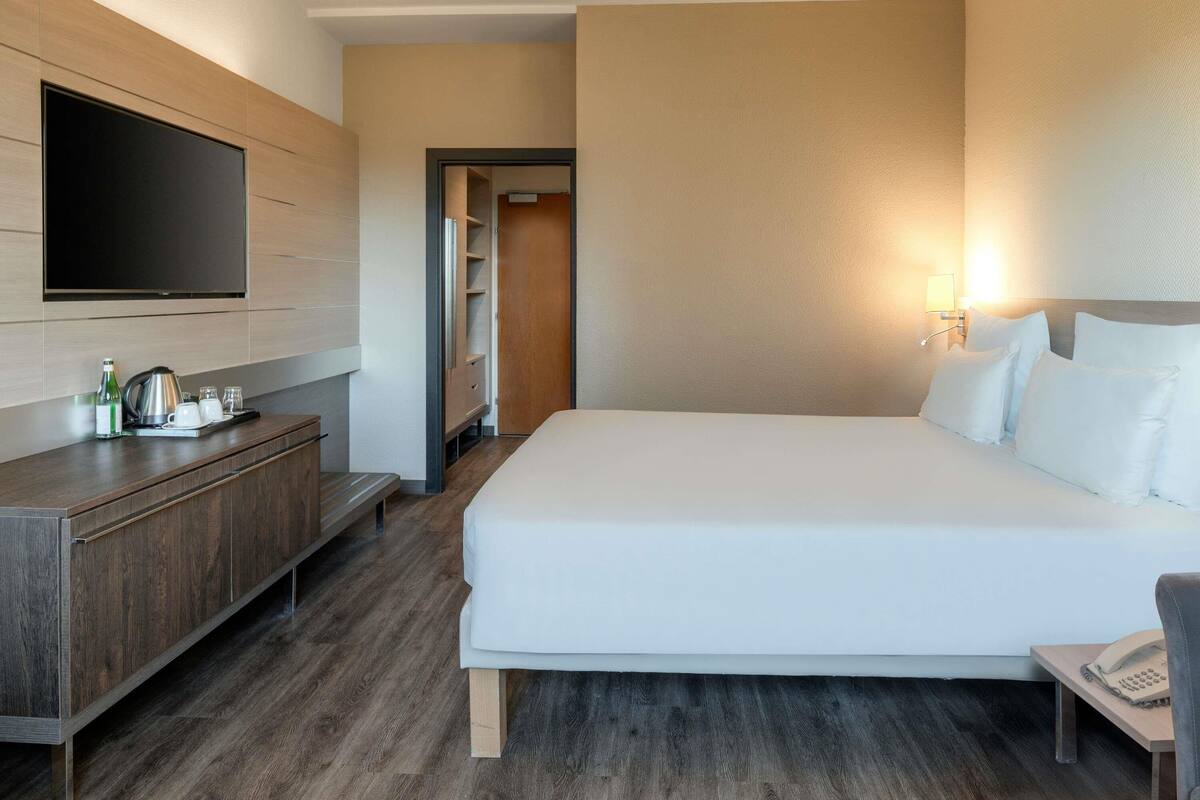 Une chambre d’hôtel moderne dotée d’un grand lit, d’un téléviseur à écran plat fixé au mur, d’une commode en bois et d’une table de chevet avec un téléphone.