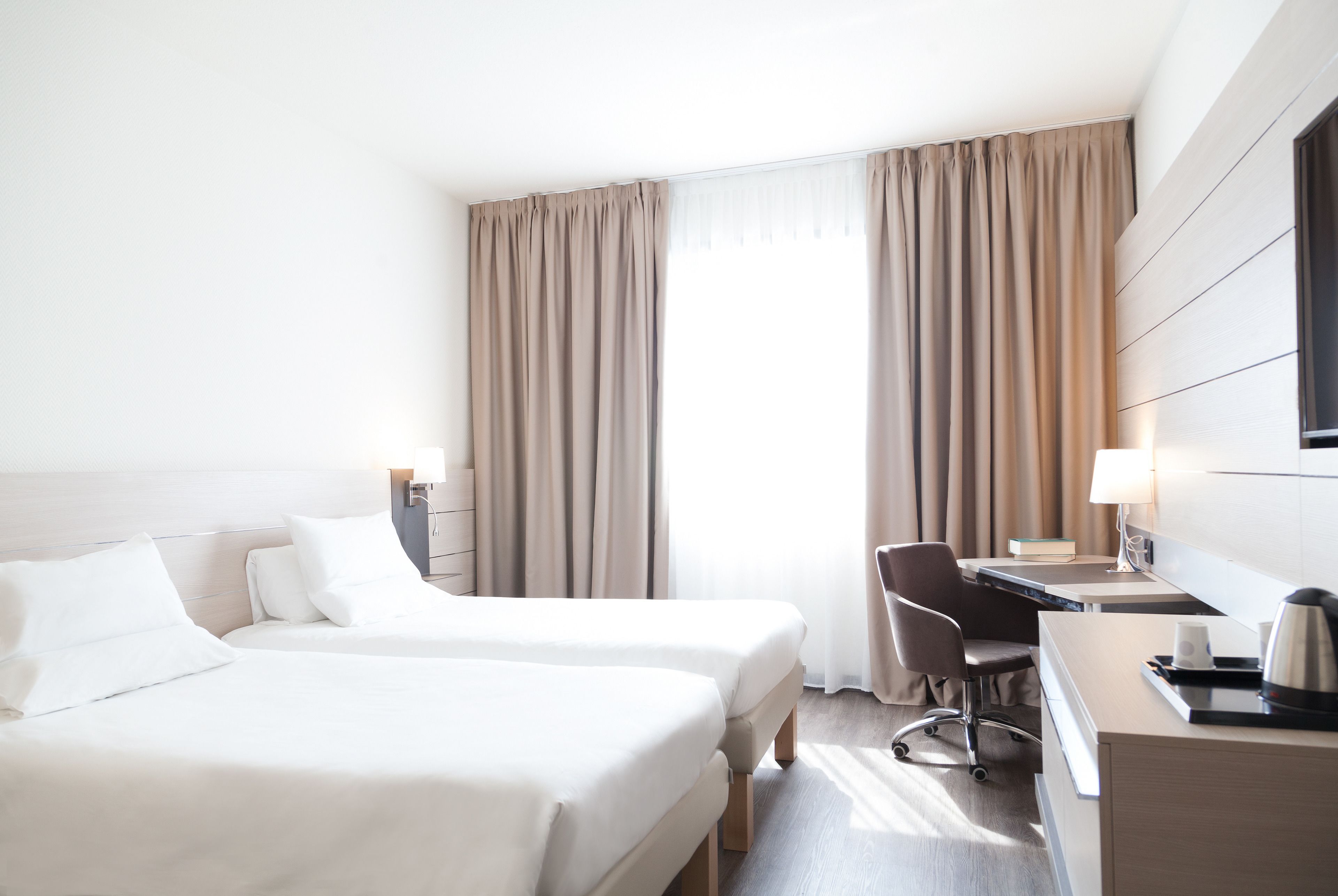 Et hotelværelse med to senge, et skrivebord, en stol, en lampe og et vindue med gardiner.