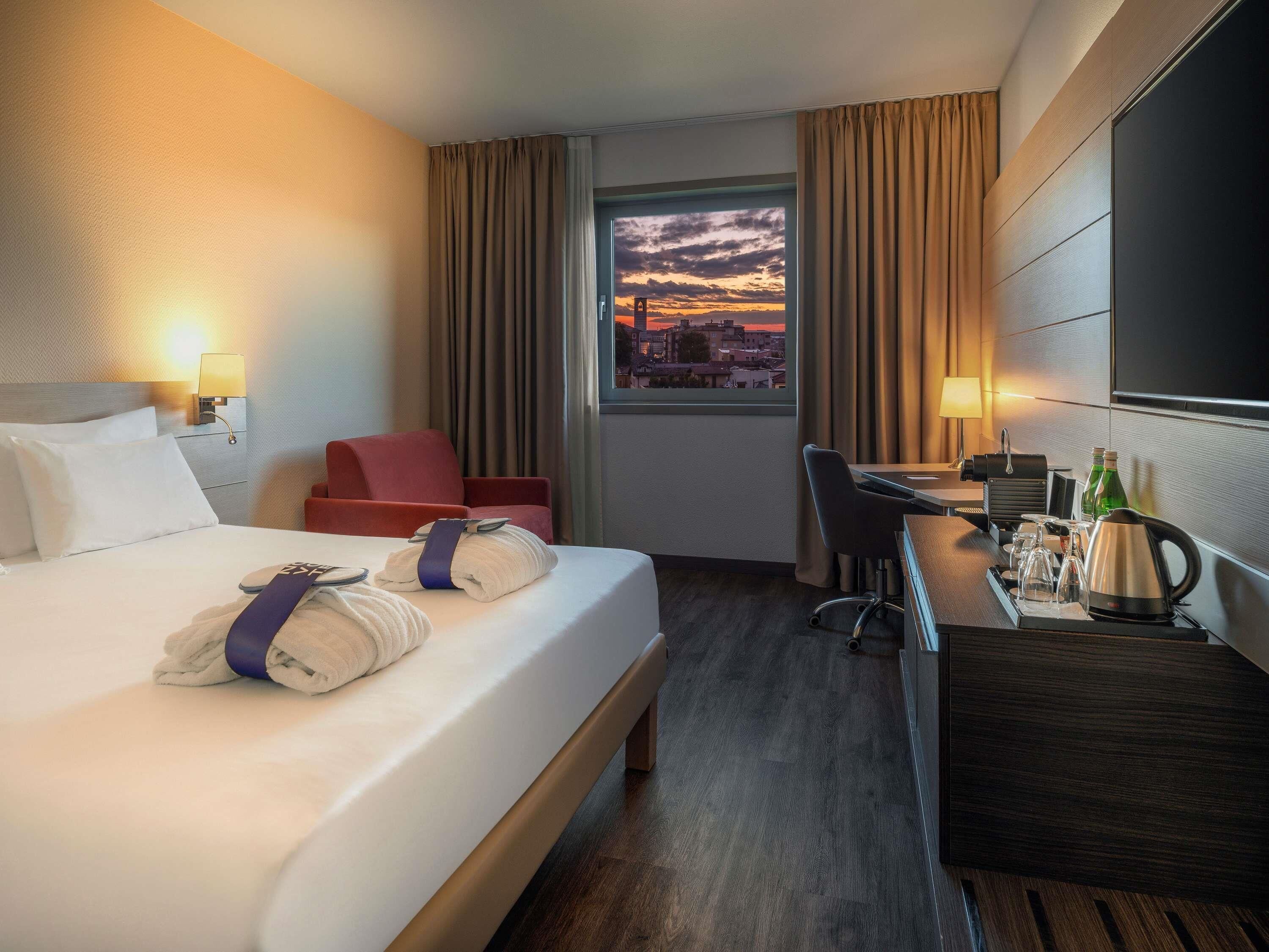 A hotel room with a bed, a desk, a chair, a window with a sunset view, and a TV on the wall.