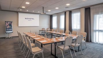 Un salón de reuniones con una larga mesa, sillas, una pantalla de proyector y un amplio ventanal con cortinas.