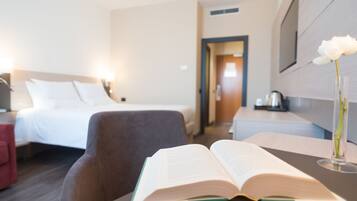 Ein Hotelzimmer mit einem Bett, einem Schreibtisch mit einem Buch, einem Sessel und einer Vase mit einer Blume.