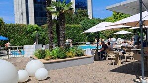 An outdoor dining area with tables and umbrellas, a swimming pool, and palm trees. - Novotel Brescia 2 (Brescia)