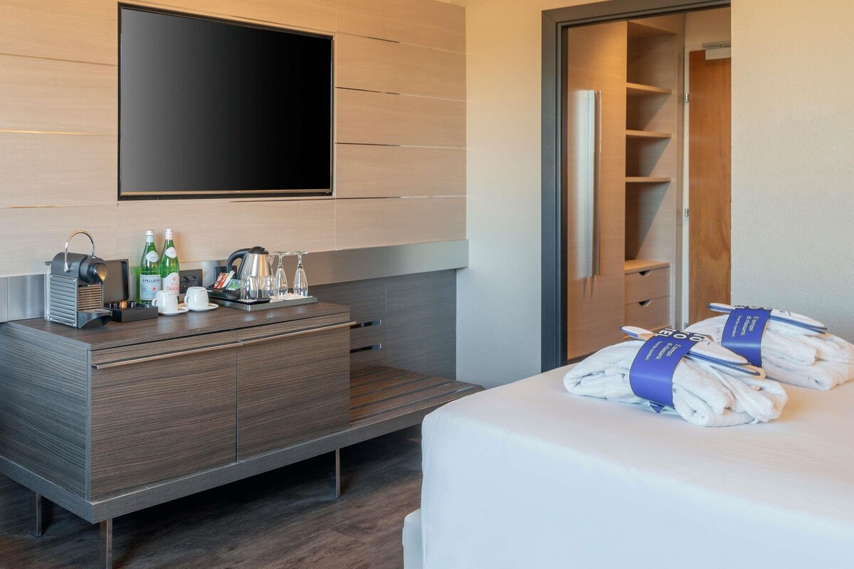 A hotel room with a flat-screen TV mounted on the wall, a wooden console table with a kettle, glasses, and bottles, a bed with towels, and a wardrobe with shelves.