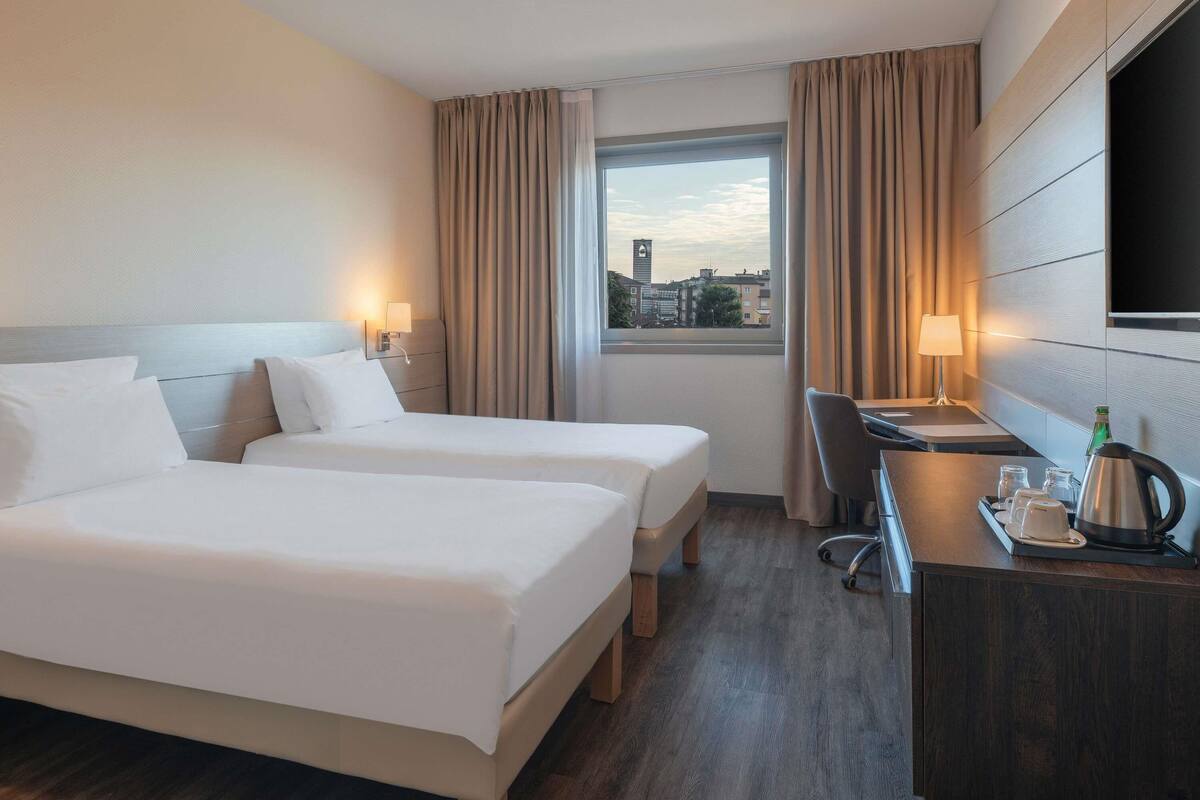 Une chambre d’hôtel avec deux lits, un bureau, une chaise, une télévision et une fenêtre avec des rideaux.