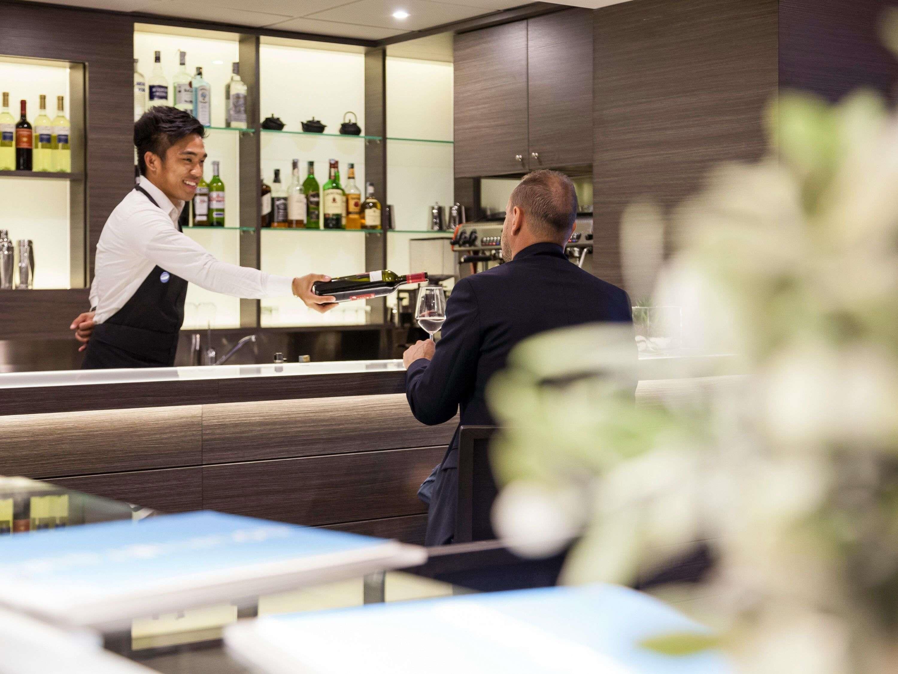 Eine Bar mit einem Barkeeper, der einen Gast bedient. Regale mit verschiedenen Flaschen und eine Theke sind ebenfalls vorhanden.