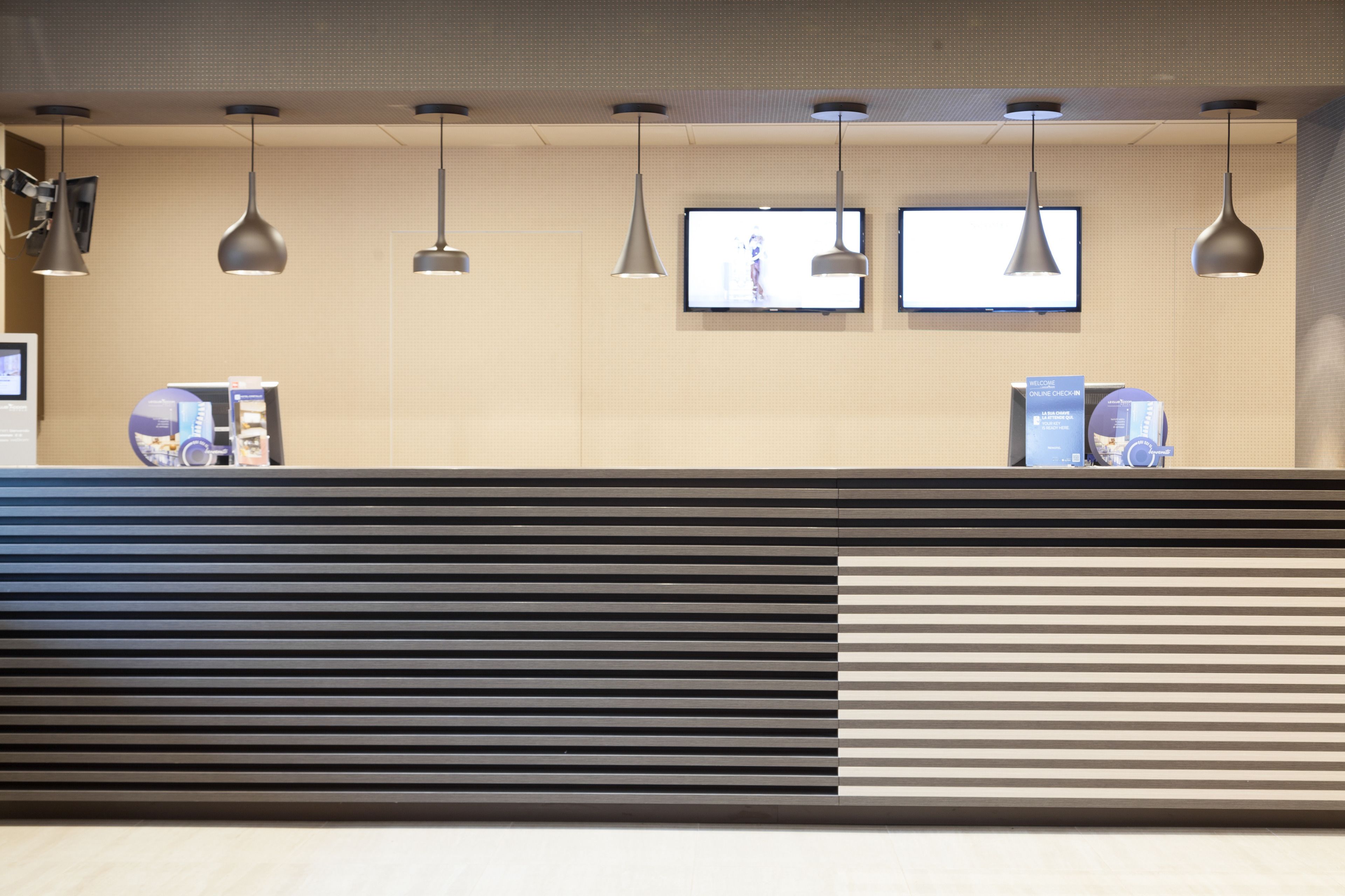 A reception desk with a modern design, featuring a black and white striped pattern, multiple pendant lights, and two flat-screen TVs mounted on the wall.