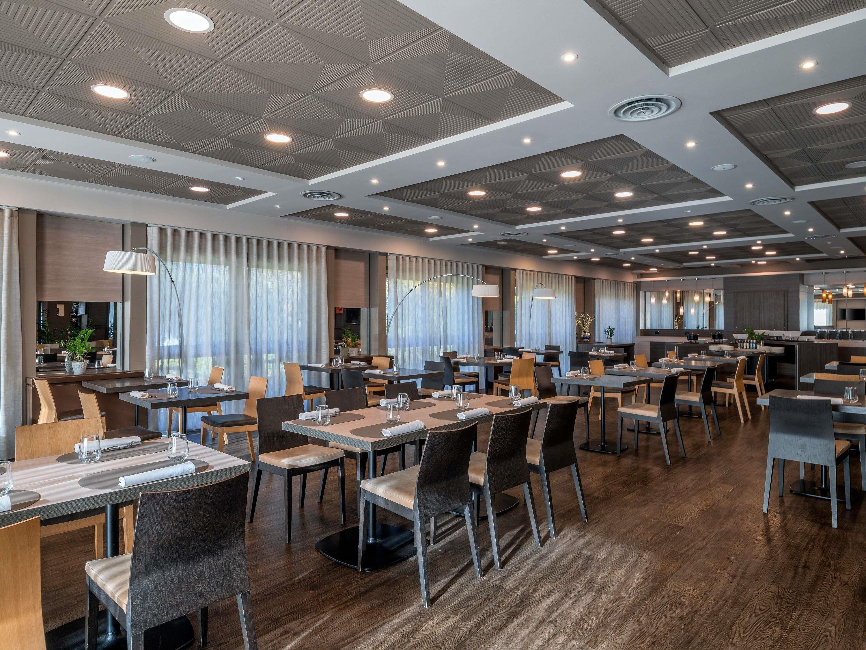 Un restaurante moderno con varias mesas y sillas, una zona de cocina e iluminación empotrada en el techo.