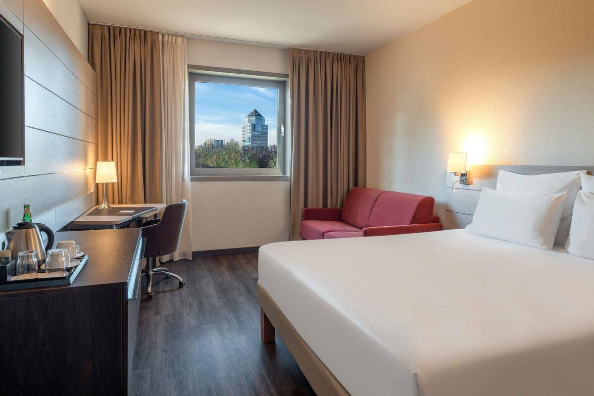 Une chambre d’hôtel comprenant un lit, un bureau, une chaise, une lampe, une fenêtre avec des rideaux et une vue sur des immeubles.