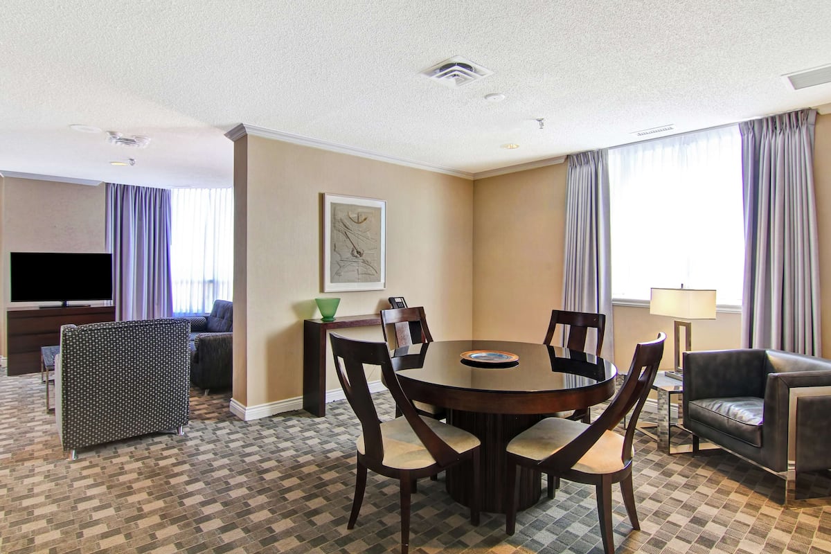 A hotel room with a dining table, chairs, a sofa, a TV, and a painting on the wall.