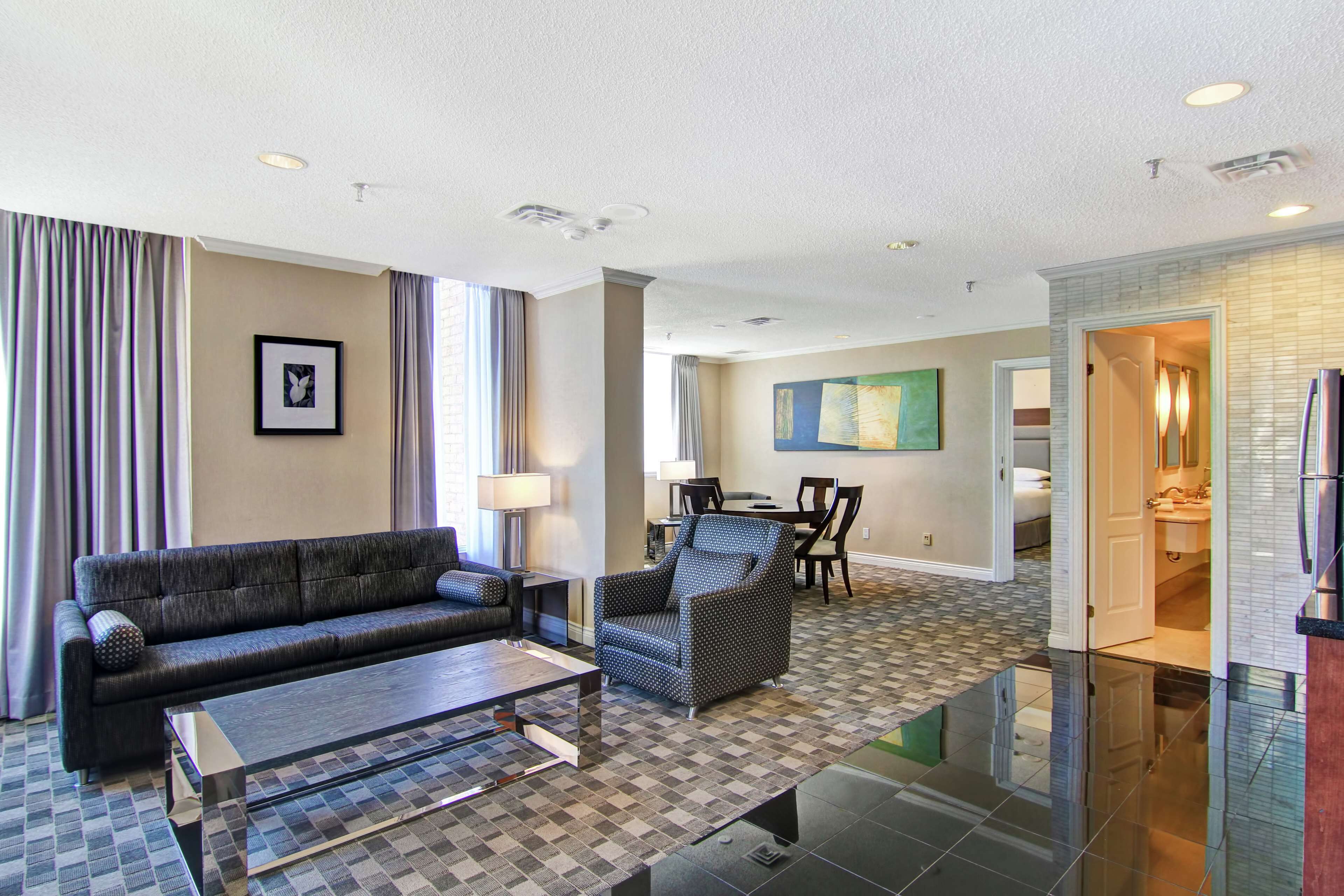 A modern hotel room with a checkered floor, a sofa, a coffee table, and a dining area with chairs and a table.