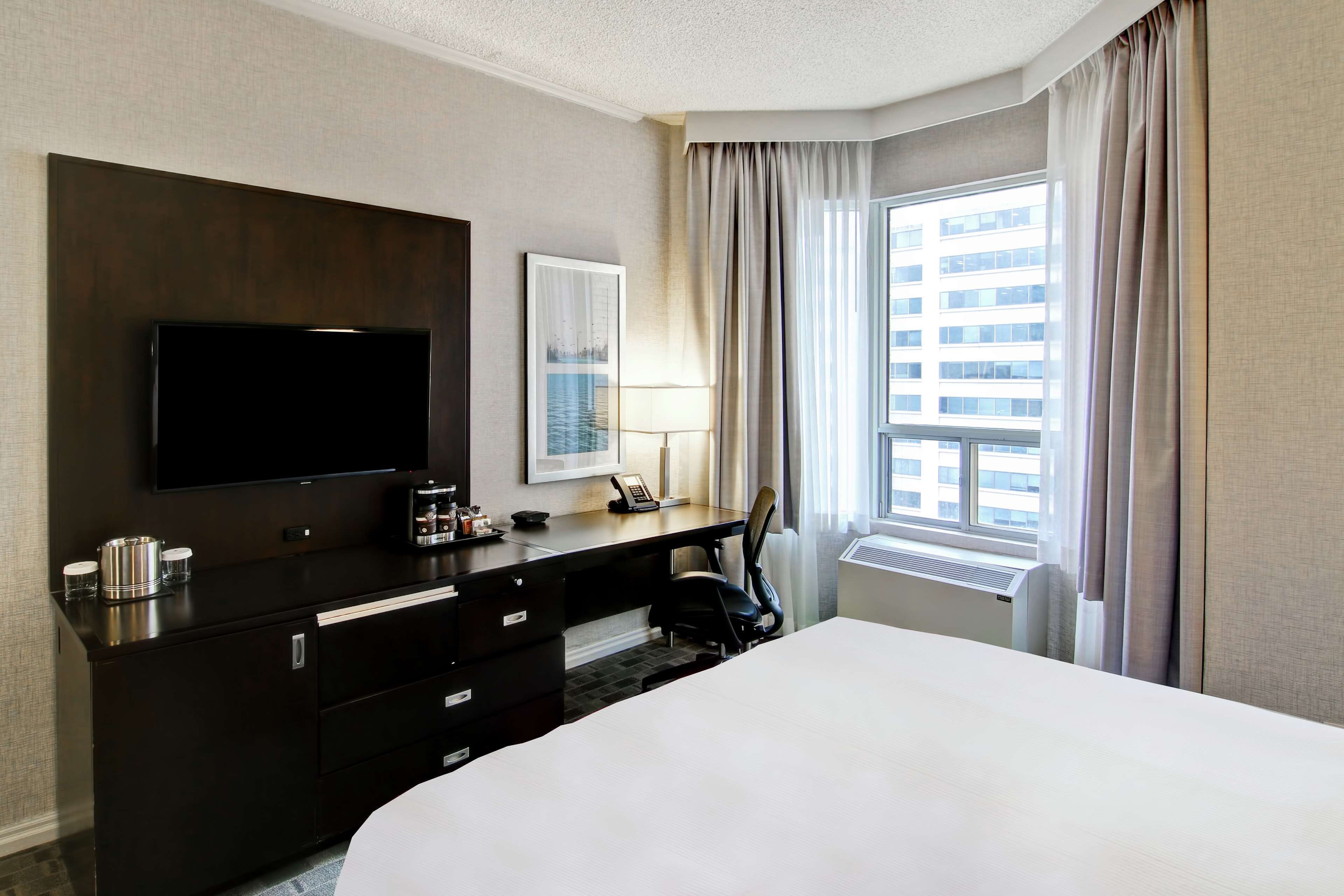 A hotel room with a large bed, a flat-screen TV mounted on the wall, a desk with a lamp, and a window with curtains.