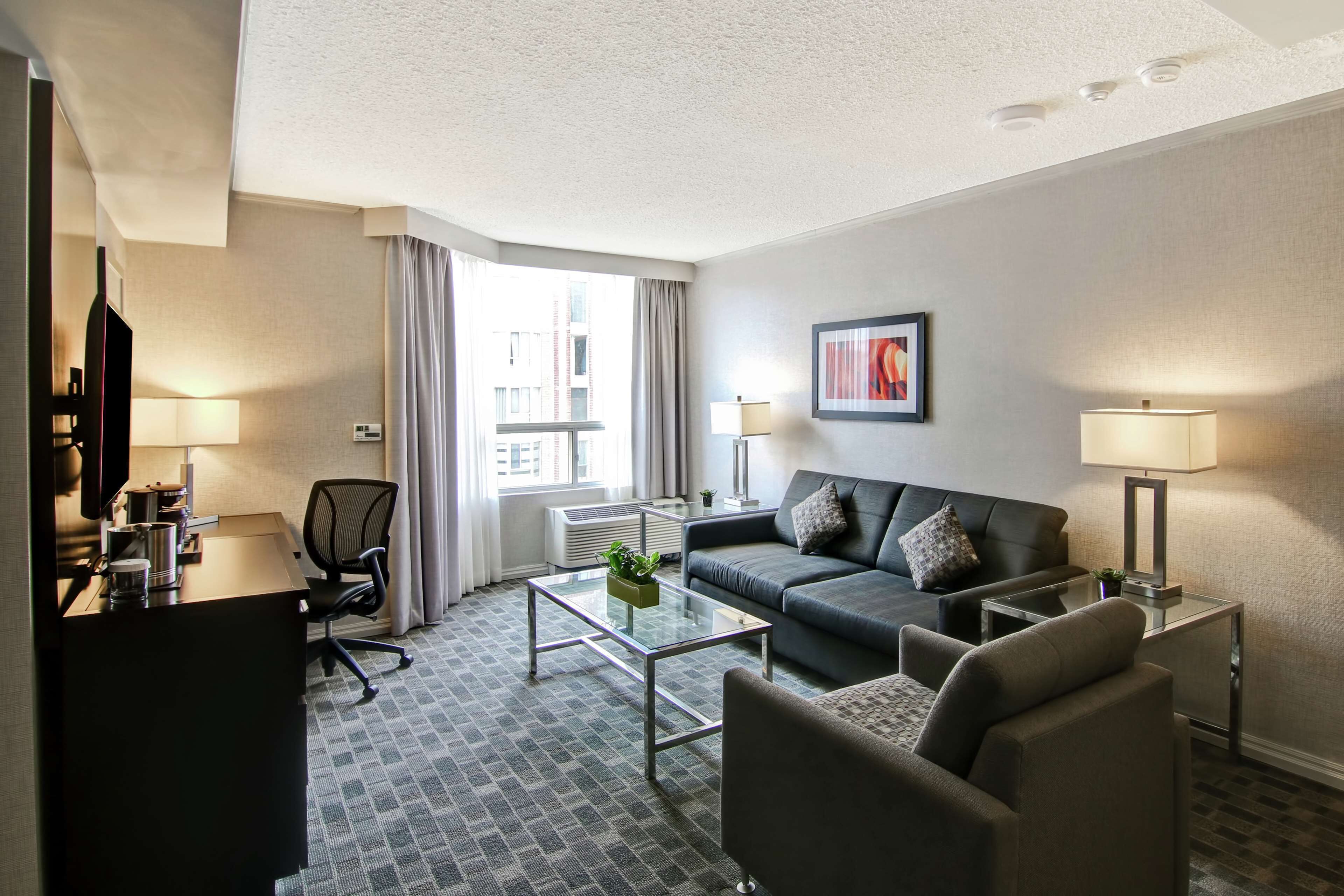 A modern hotel room with a dark grey sofa, a glass coffee table, a desk with a computer, and a large window with curtains.