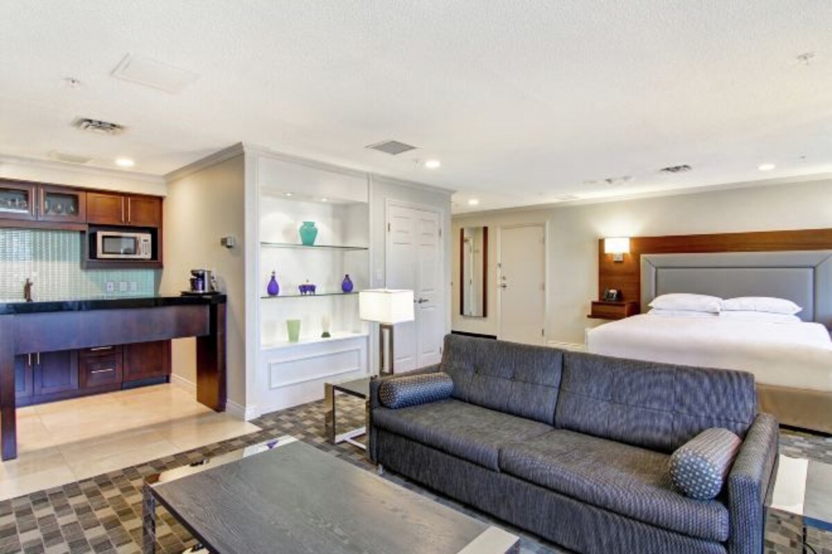 Una habitación de hotel moderna con kitchenette, un sofá gris, una cama y una mesita de noche con lámpara.
