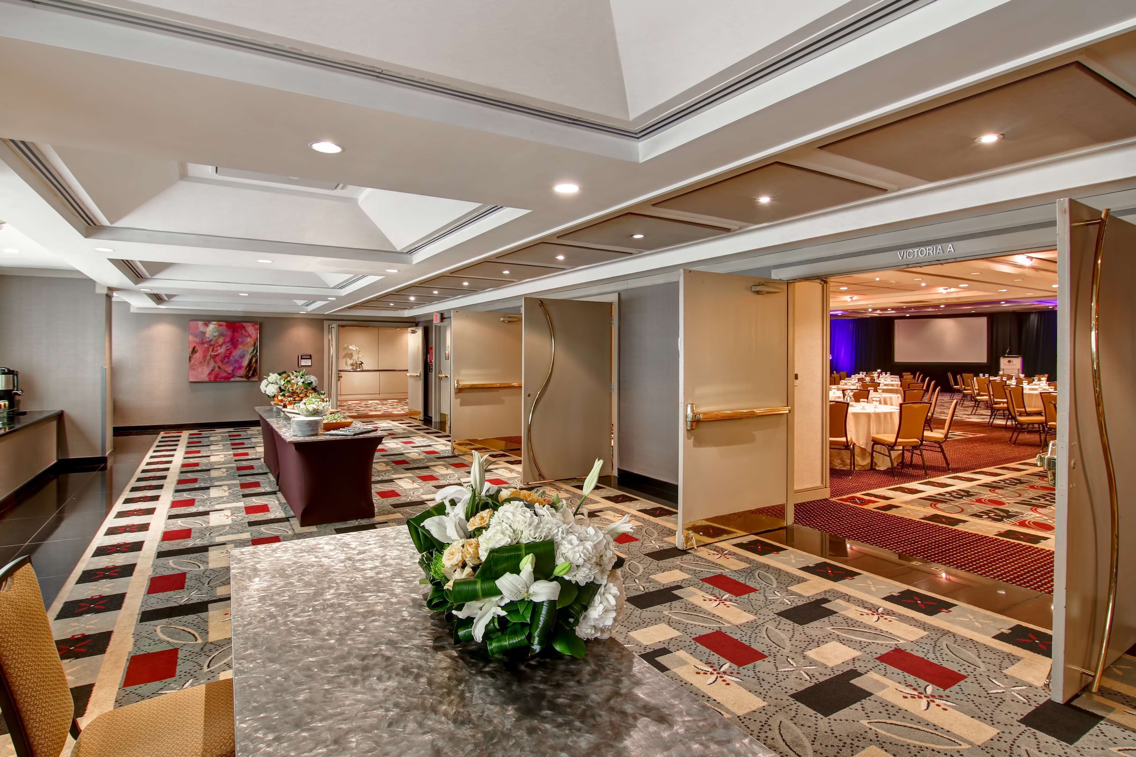 Et hotel lobby med en receptionsdisk, en blomsteropsætning på et bord, et mønstret tæppe og et mødelokale, der kan ses igennem en åben dør.