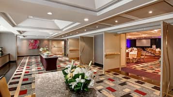 Un hall de hotel con mostrador de recepción, un centro de mesa floral sobre una mesa, alfombra estampada y una sala de conferencias visible a través de una puerta abierta.