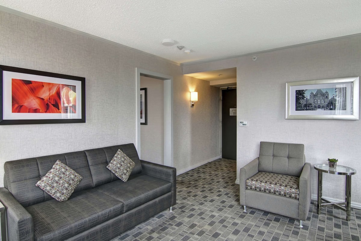 A hotel room with a grey sofa, a grey armchair, a small round table with a plant, and two framed pictures on the walls.