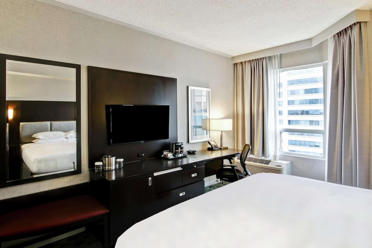 A hotel room with a large bed, a flat-screen TV, a desk with a lamp, a telephone, and a window with curtains.