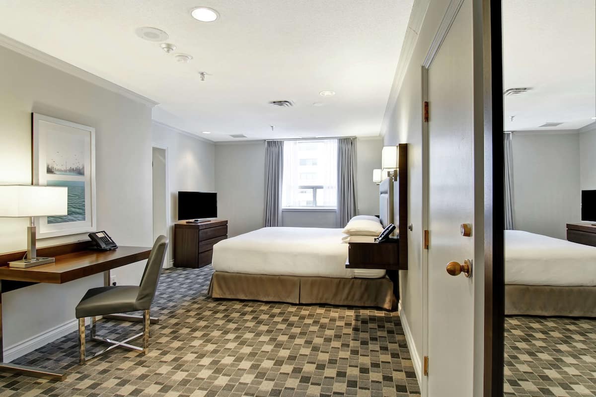 A hotel room with a bed, desk, chair, and a view of the city through the window.