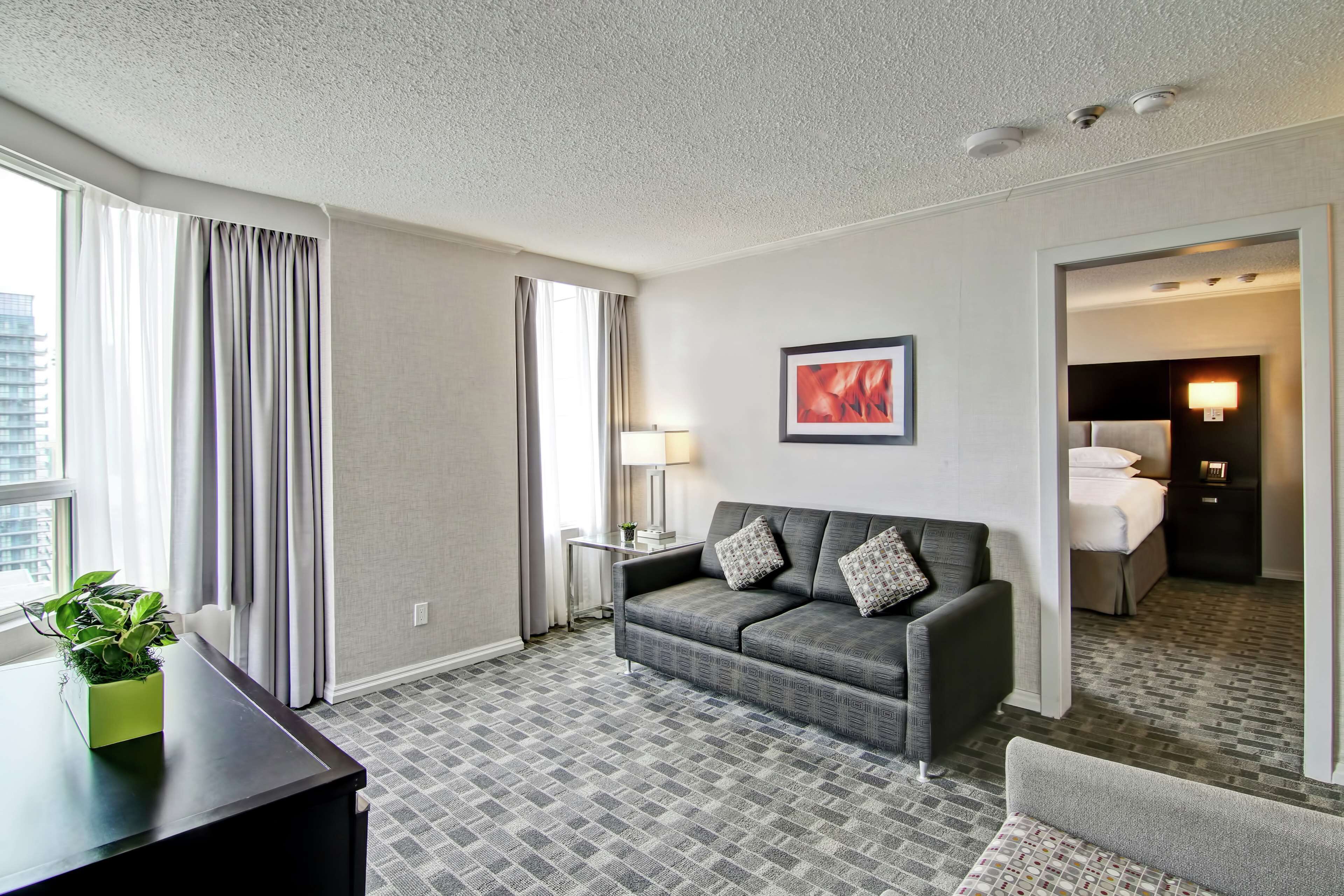 Quarto de hotel com carpete cinza estampado, sofá cinza com almofadas padronadas, mesa pequena com abajur, quadro pendurado na parede e vista da cidade pela janela.