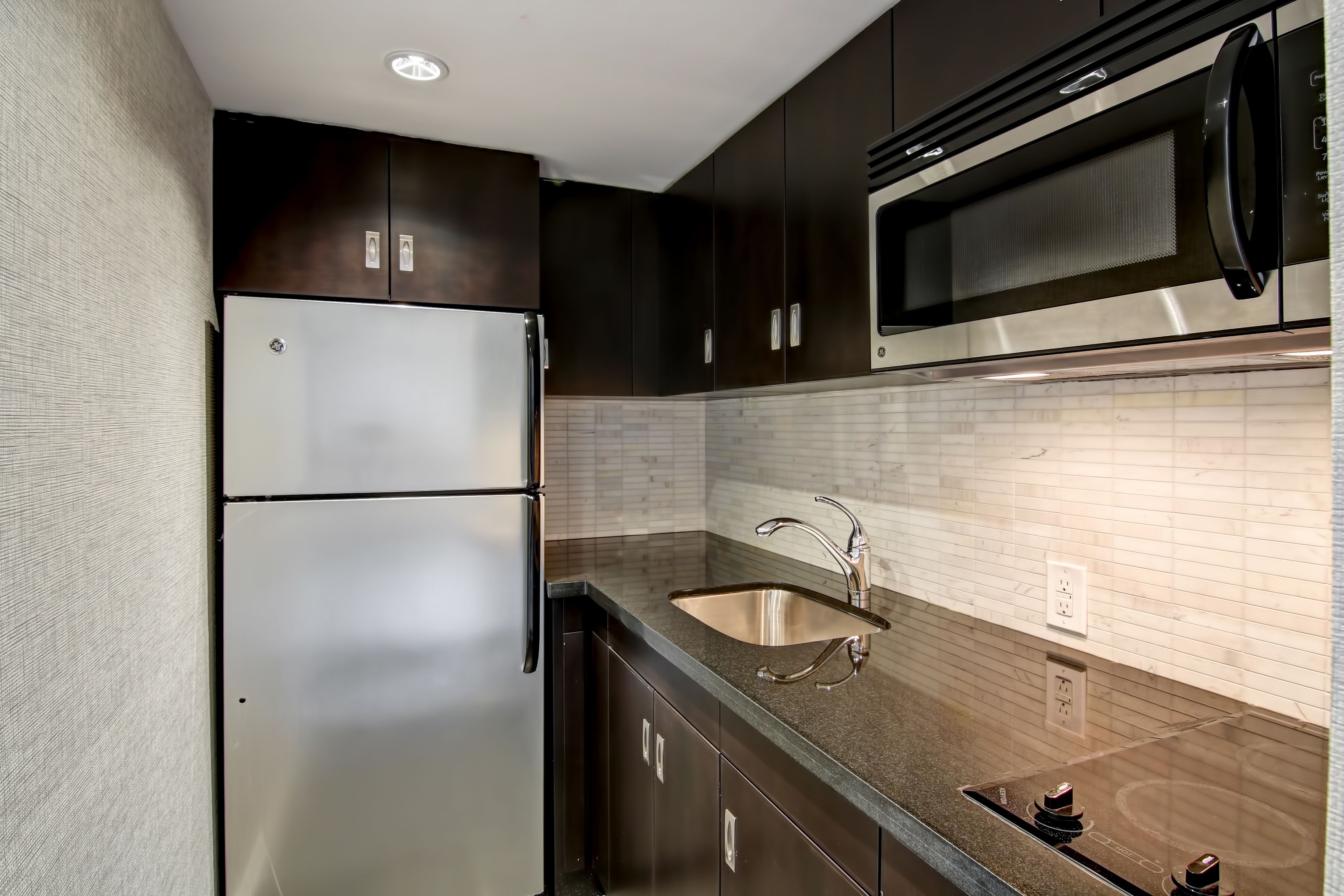 A compact kitchen with dark cabinetry, a stainless steel refrigerator, a microwave, and a sink with a modern faucet.