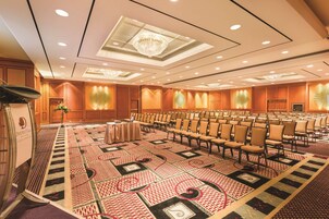 Een ruime congreszaal met rijen stoelen, een spreekgestoelte en een kamerbreed tapijt met dessin.